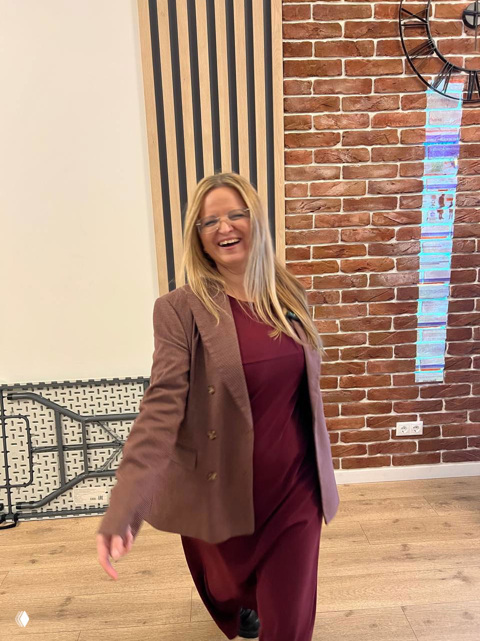 Женщина в пиджаке на фоне кирпичной стены и большого настенного часов, смеётся в офисной обстановке на деревянном полу.