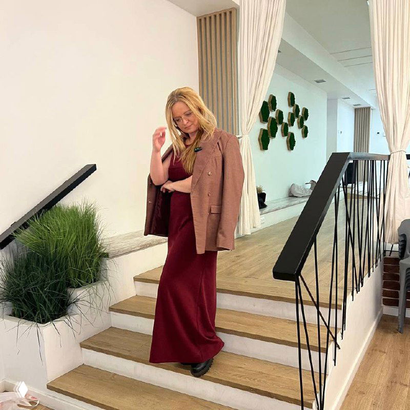 Женщина в бордовом платье и пиджаке спускается по деревянной лестнице внутри светлого офиса, взгляд направлен вниз.