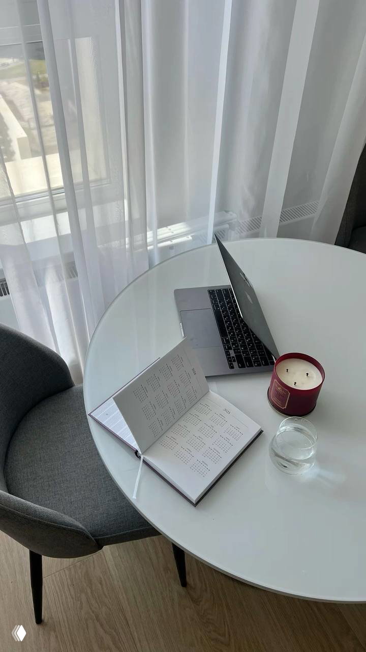 Круглый белый стол у окна с открытым ноутбуком, планером и свечой — сценка для планирования, учёта и подготовки годовой отчётности.