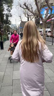 Вид сзади женщины в длинном светлом пальто при прогулке по улице Шанхая среди пешеходов и городской среды, прохожие и уличная суета вокруг.