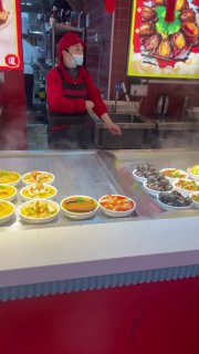 Прилавок уличной еды в Китае: керамические миски с различными горячими блюдами, повар в маске готовит за стеклом на кухне фудкорта, уличная гастрономия.