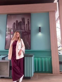 Женщина в бордовом платье и светлом пальто стоит в кафе на фоне постера с небоскрёбами; интерьер с зелёной отделкой и декоративной панелью.