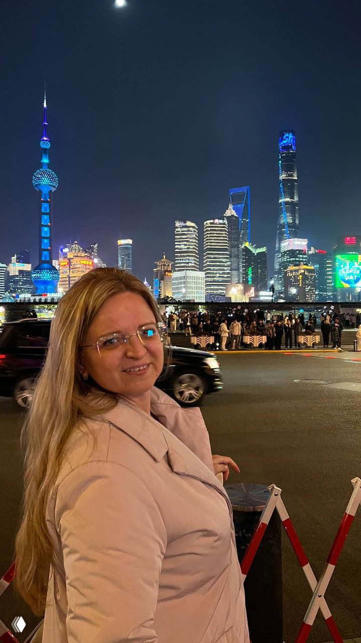 Портрет автора/путешественницы на набережной с панорамой Путона и ярко подсвеченными небоскрёбами Шанхая на заднем плане, вечерняя прогулка туристов.