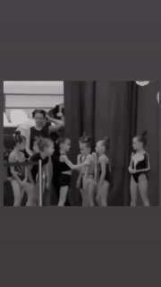 Чёрно‑белый кадр с группой юных гимнасток в спортивных купальниках за кулисами: одна девочка получает объятие от подруг перед выходом.
