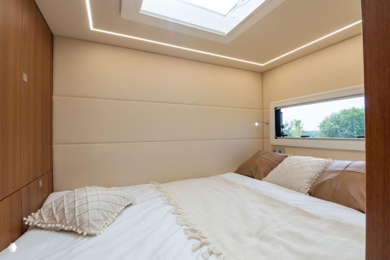 Спальная зона автодома с широкой кроватью, панорамным люком в потолке и мягкой обшивкой стен для комфортного отдыха в дороге.