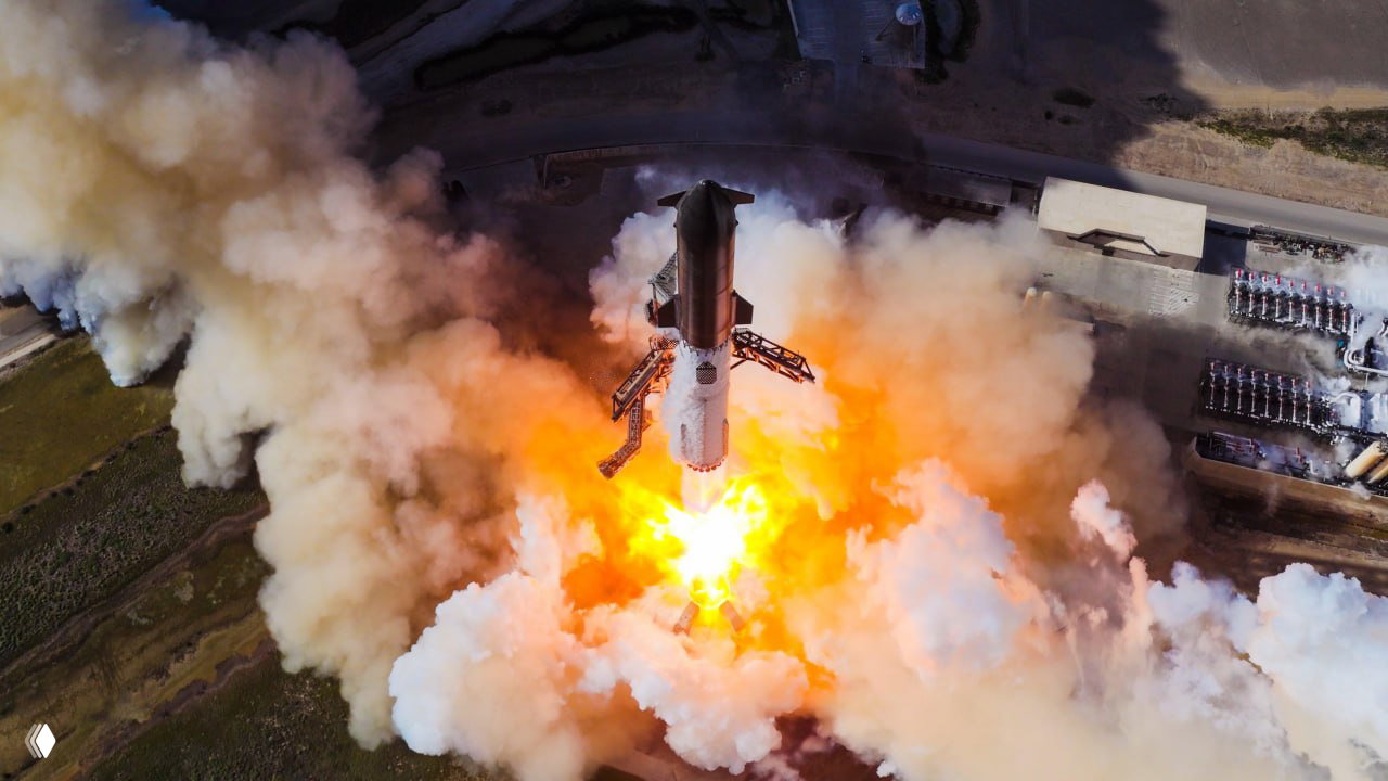 Аэрофотосъёмка старта Starship сверху: ракета на вертикали, вокруг — огромные облака пыли и дыма, видна стартовая инфраструктура рядом.