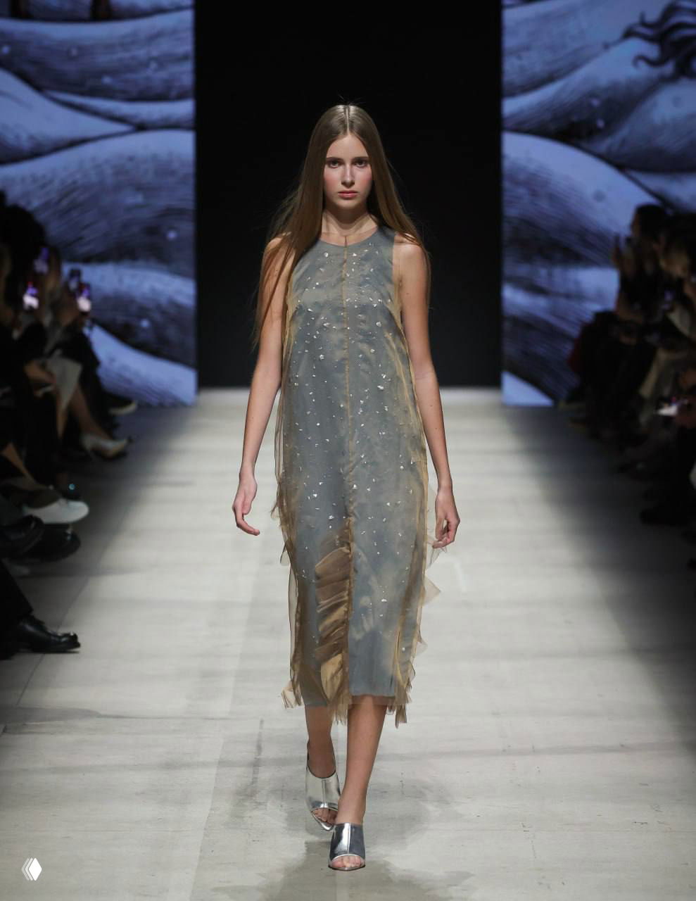 Модель в полупрозрачном серо-голубом платье с декоративной вышивкой идёт по подиуму показа студентов Сергея Сысоева.