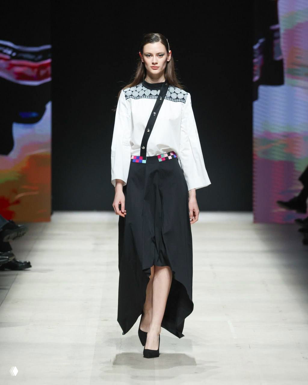 Модель в белой блузе с контрастной отделкой и асимметричной чёрной юбкой дефилирует по подиуму во время шоу Московской недели моды в Манежe.