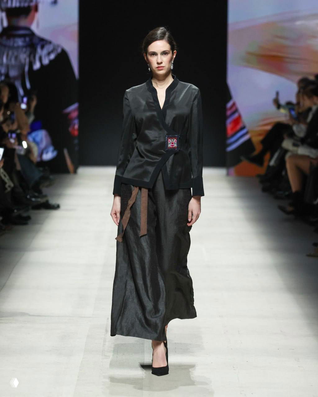 Модель в строгом чёрном костюме с длинной юбкой идёт по подиуму, подчёркнуты линии кроя и минималистичный силуэт на Московской неделе моды.