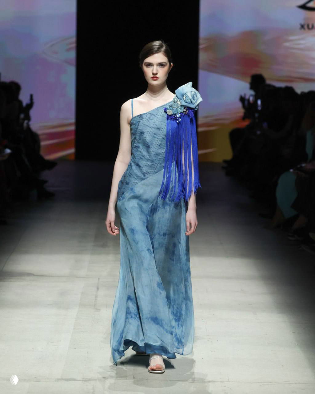 Модель в голубом асимметричном платье с декоративной кистью на плече дефилирует по подиуму на шоу, цвет и текстура платья на фоне сцены.
