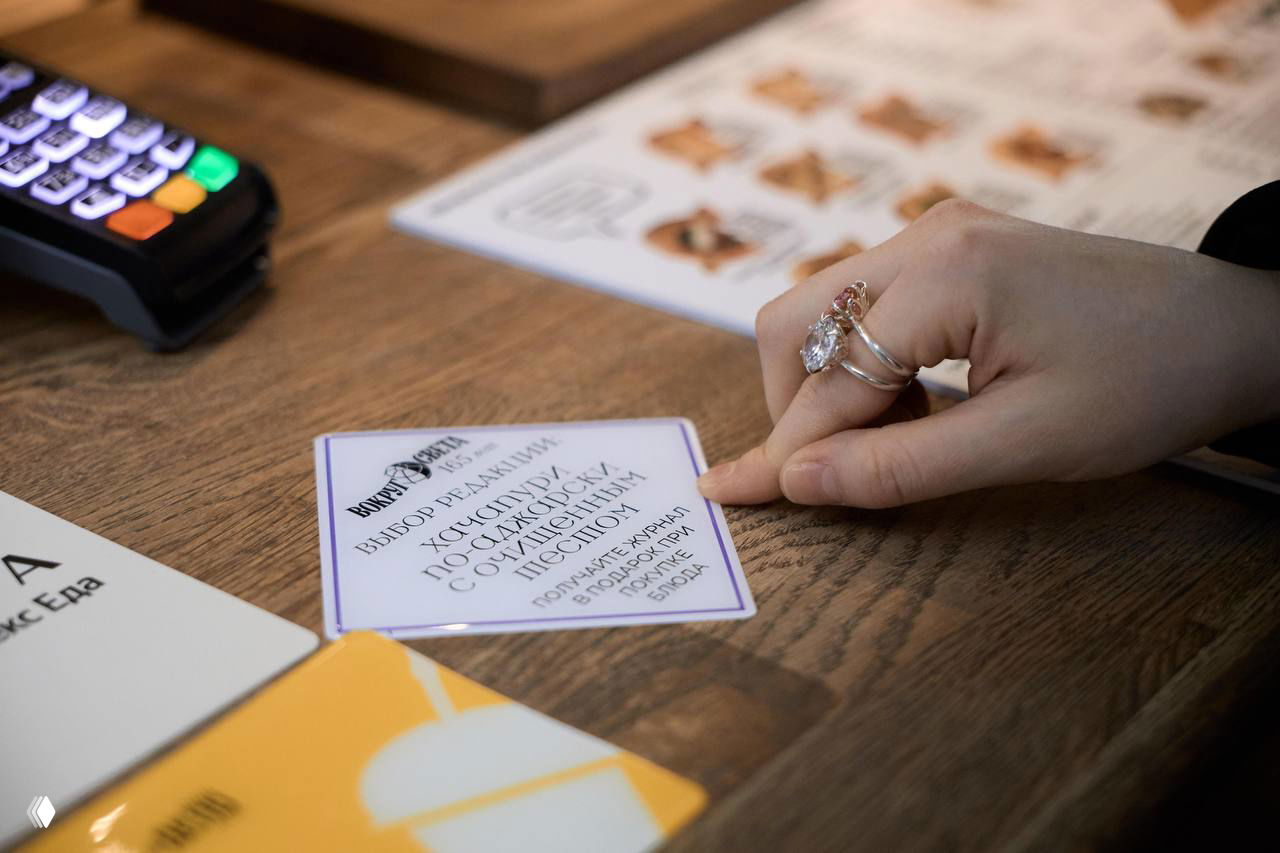 Рука участника у стола держит карточку «Выбор редакции» рядом с меню и терминалом оплаты, демонстрируя гастрономические позиции фестиваля в корнерах ДЕПО.