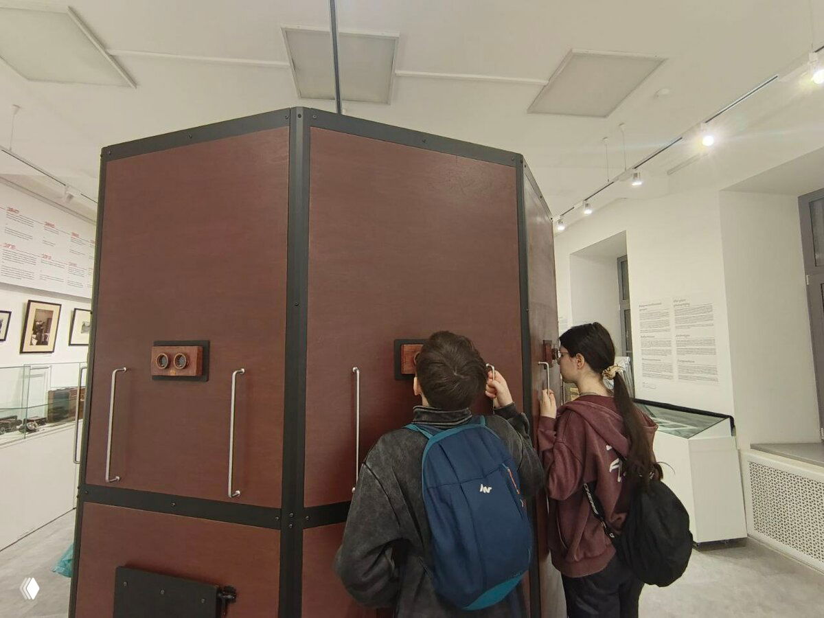 Дети в рюкзаках рассматривают экспонат в музее: двое детей у крупного деревянного интерактивного стенда с окулярами и смотровыми окнами.