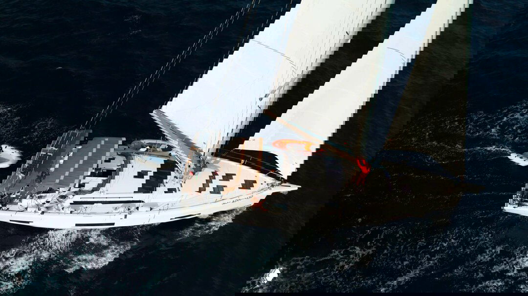 Аэрофотосъёмка парусной яхты в движении: вид на кокпит, тент и пассажиров, подчёркнуты белые паруса и след от борта на воде.