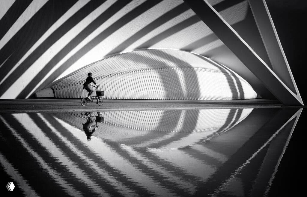 Чёрно‑белый кадр с велосипедистом и архитектурными тенью и отражением в воде; сильная графическая композиция — финалист LCE 2026.