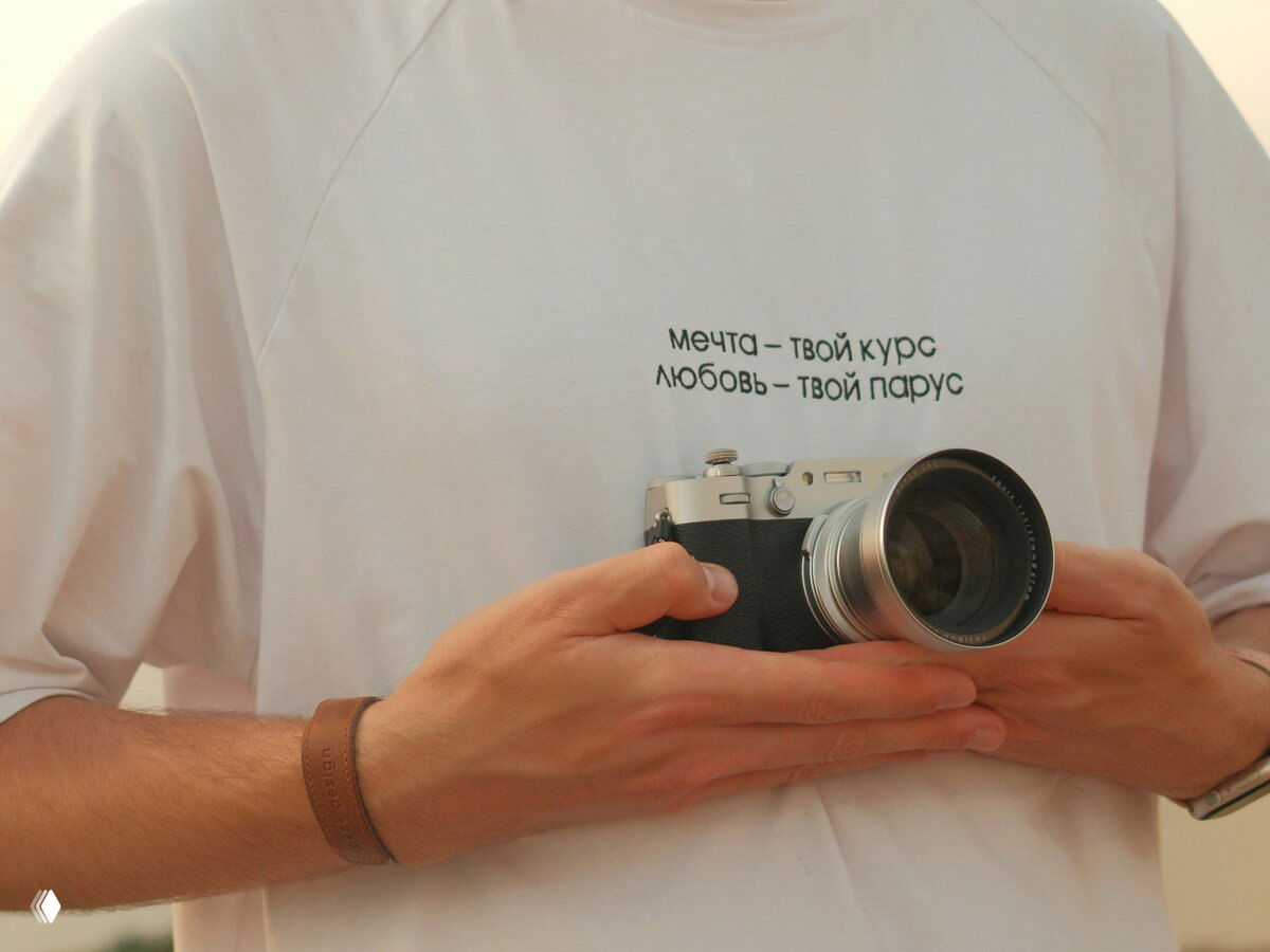 Руки человека, держащие фотокамеру; на футболке видна надпись «мечта — твой курс любовь — твой парус», подчёркивающая романтику путешествия.