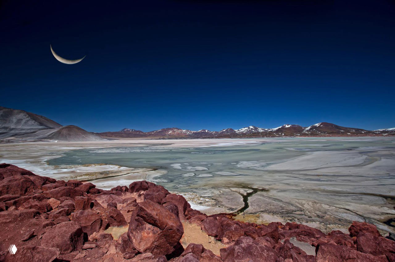 Пейзаж с луной над солончаком и горными отрогами: освещённые скалы и пресноводные заливы создают причудливую палитру текстур и тонов на переднем плане.