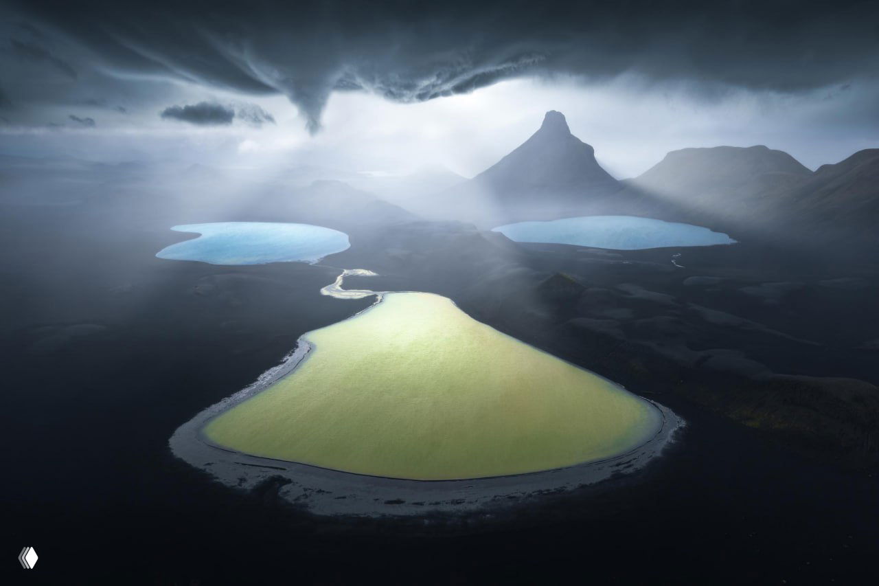 Мрачный атмосферный пейзаж с озёрами необычного цвета и силуэтом гор в тумане: драматичное освещение подчёркивает рельеф и формы ландшафта.