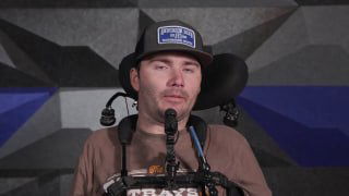 Мужчина в кресле с опорой для головы и микрофоном, на фоне оборудование для демонстрации нейроимпланта и роботизированной руки во время презентации