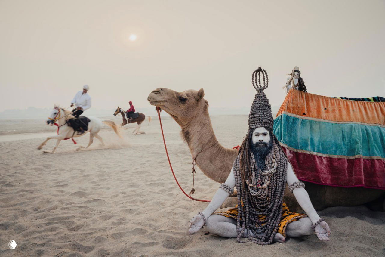 Уличный сюжет на песчаном побережье: верблюд и человек в традиционной одежде на переднем плане, на заднем плане скачут всадники — кадр из серии «Путешествия».
