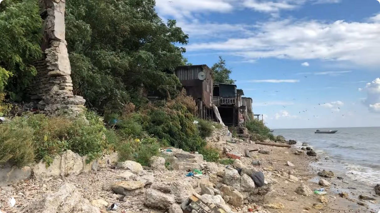 Пляж с валунами и обломками бетона, старые домики и пристройки над водой; лодка у горизонта и прибрежная растительность.