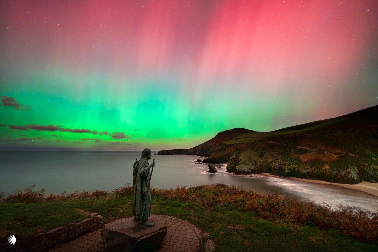 Палатки на лугу при ночном наблюдении, небо озарено ярким розово‑зелёным сиянием над тёмной линией горизонта и звёздным фоном.