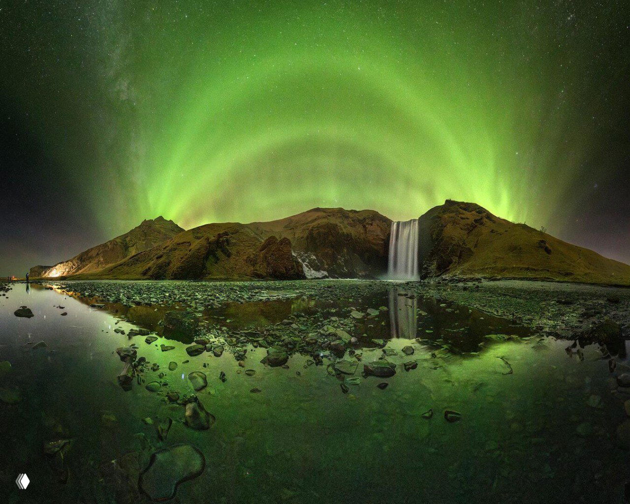 Широкоформатный кадр водопада и горного ландшафта, над которыми выстроилась симметричная зелёная арка северного сияния с отражением в лужах.