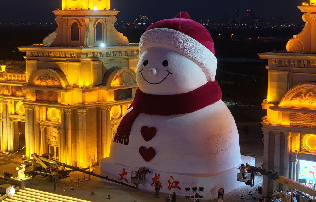 Ночная съемка подсвеченного гигантского снеговика в Харбине рядом с освещенными архитектурными башнями, видны рабочие у основания.
