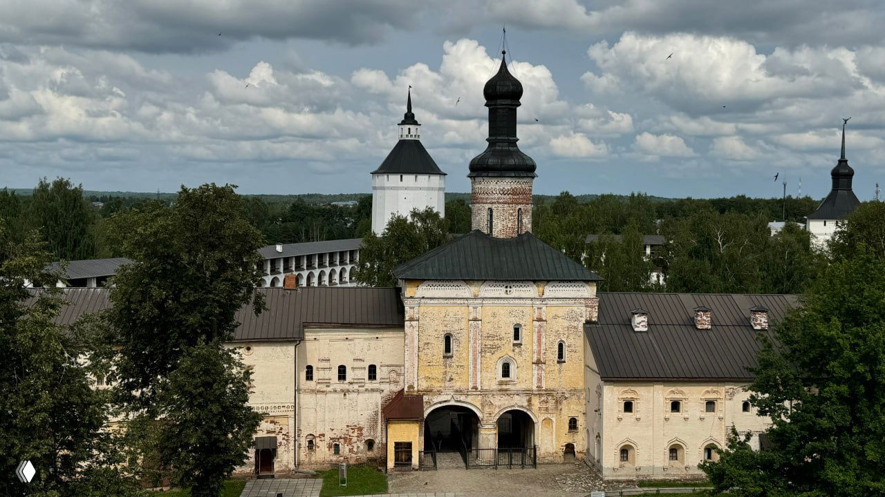 Вид на главный вход и фасады Кирилло‑Белозерского монастыря среди зелёной растительности, белые стены и характерные шатровые и луковичные купола под облачным небом.