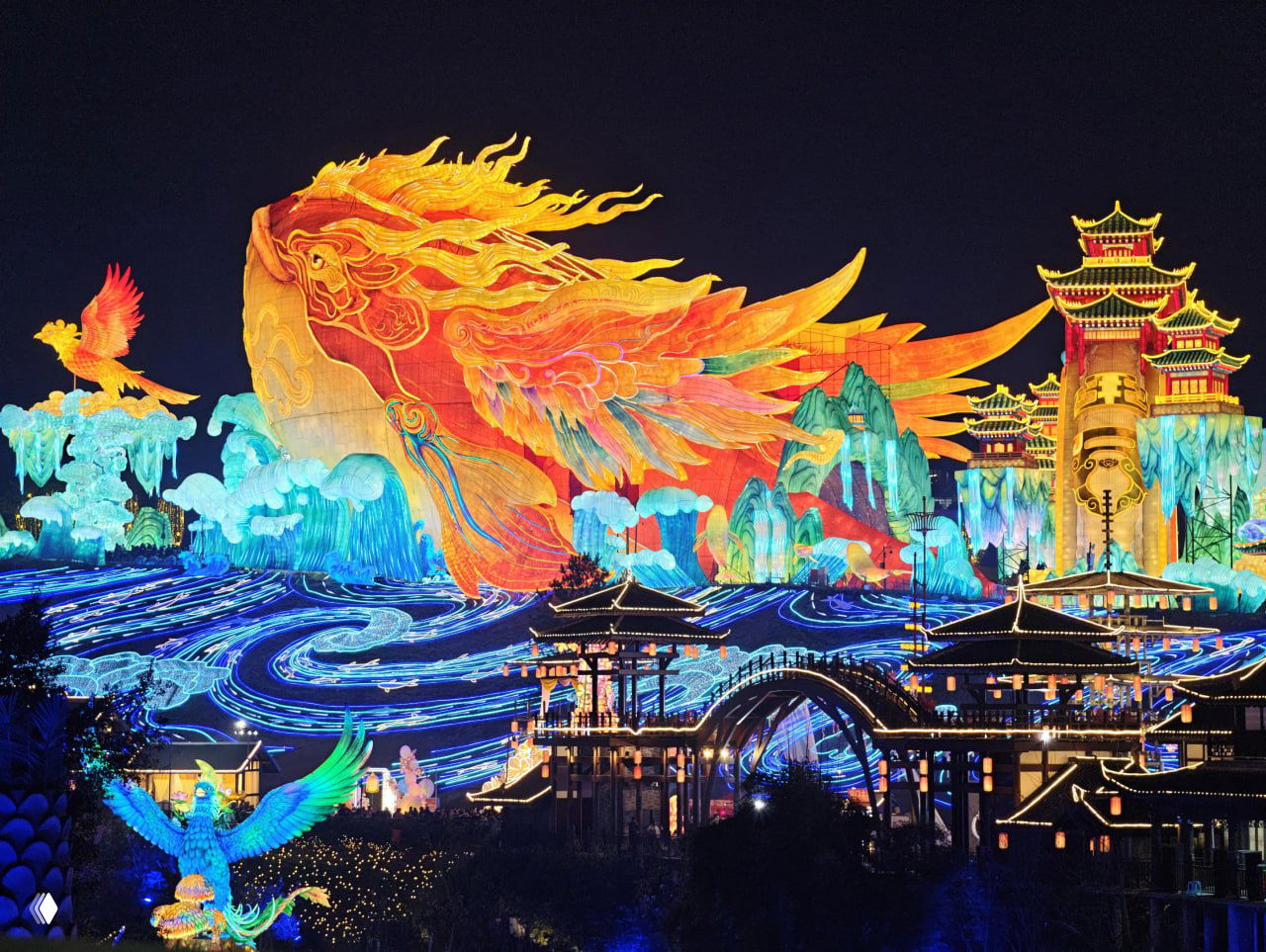 Огромная цветовая инсталляция в форме мифической птицы и пейзажа: сложная подсветка, яркие волнообразные узоры и архитектурные элементы фестиваля ночью.