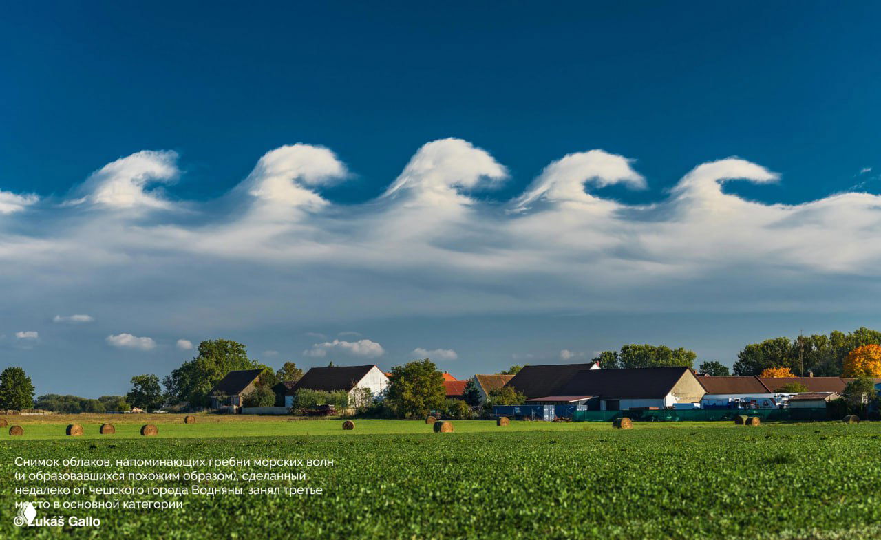 Пейзажная фотография: облака, напоминающие гребни морских волн над сельской местностью, дома и зелёное поле на переднем плане.