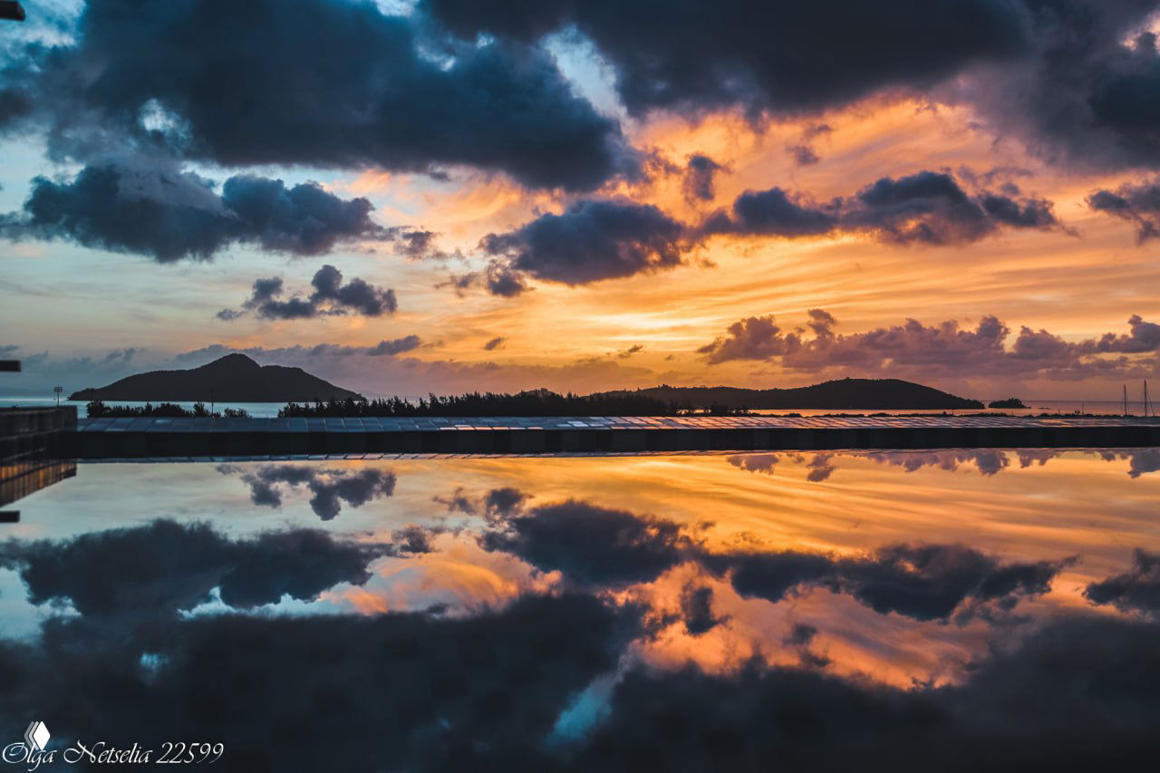 Драматичный золотисто-оранжевый закат, отражающийся в зеркальной поверхности бассейна, силуэт острова и облака, создающие живописную композицию.