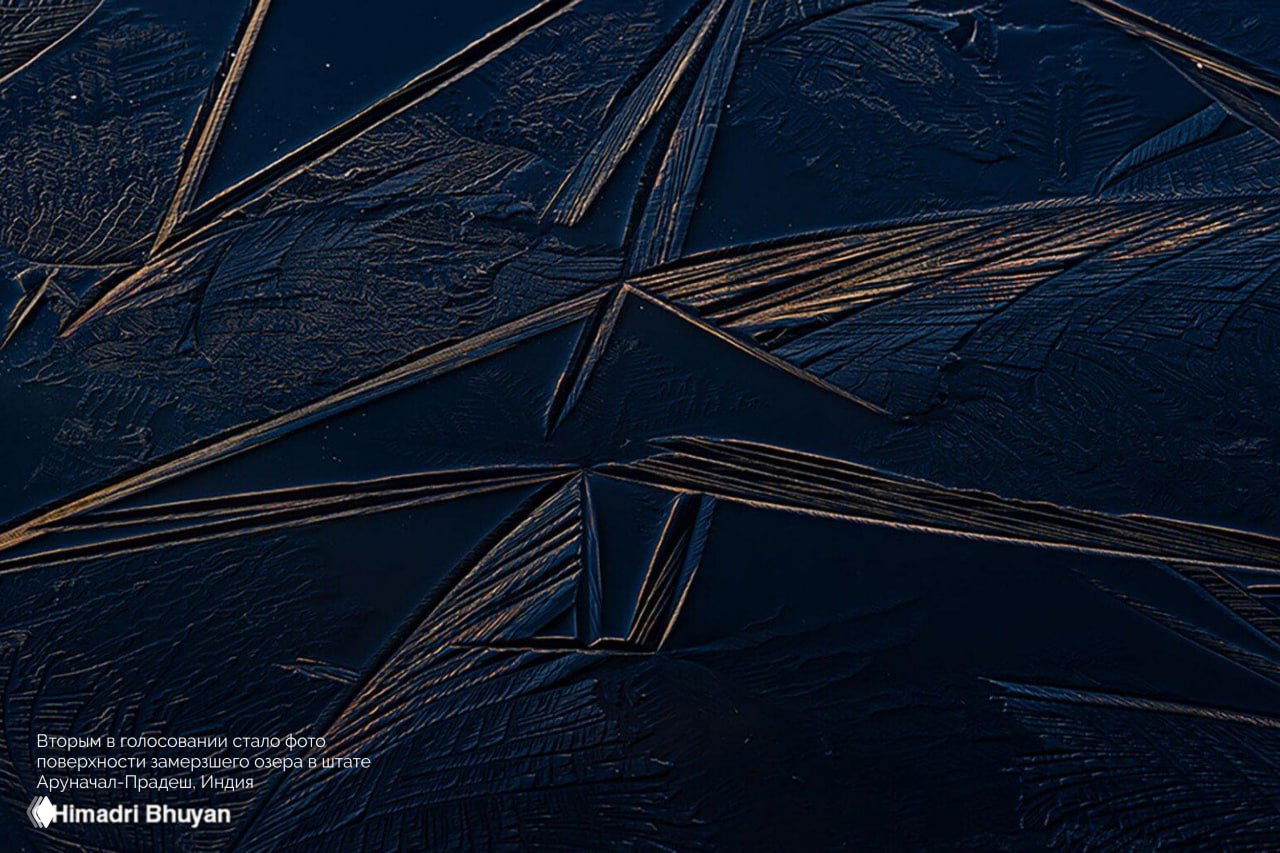 Макроснимок замёрзшей поверхности озера с геометрическими трещинами и текстурой льда, тёмная холодная тональность кадра.