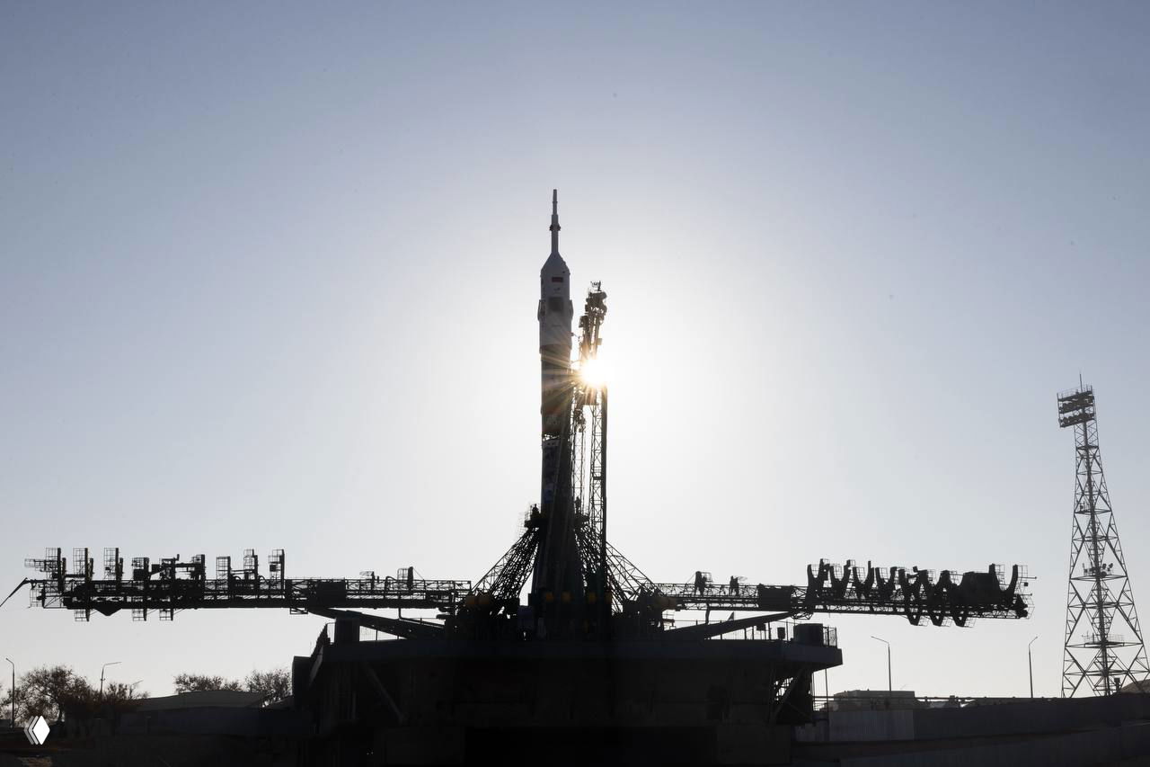Силуэт ракеты «Союз» на старте при низком солнце; контур стартового комплекса, обслуживающий персонал и подъёмные конструкции видны на фоне неба.