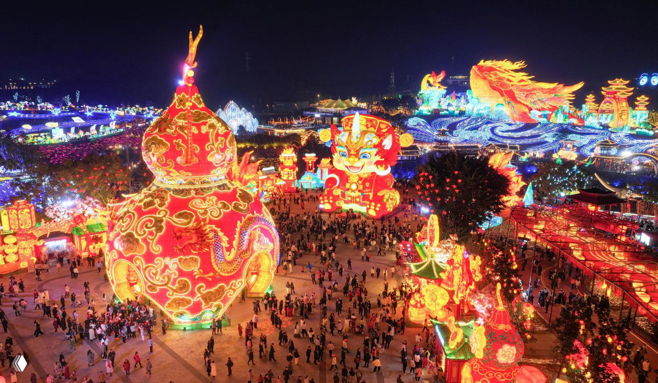 Общий вид на площадь фестиваля с большими фигурами и инсталляциями, ярко подсвеченными элементами и потоком людей, гуляющих между композициями вечером.