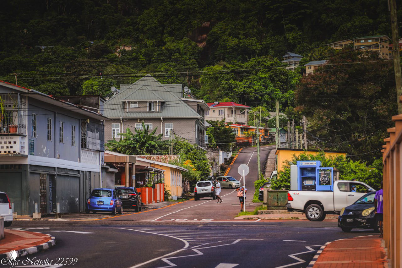 Уличная сцена сейшельской деревни с домами, припаркованными автомобилями, пешеходами и крутым зелёным холмом на заднем плане под дневным небом.
