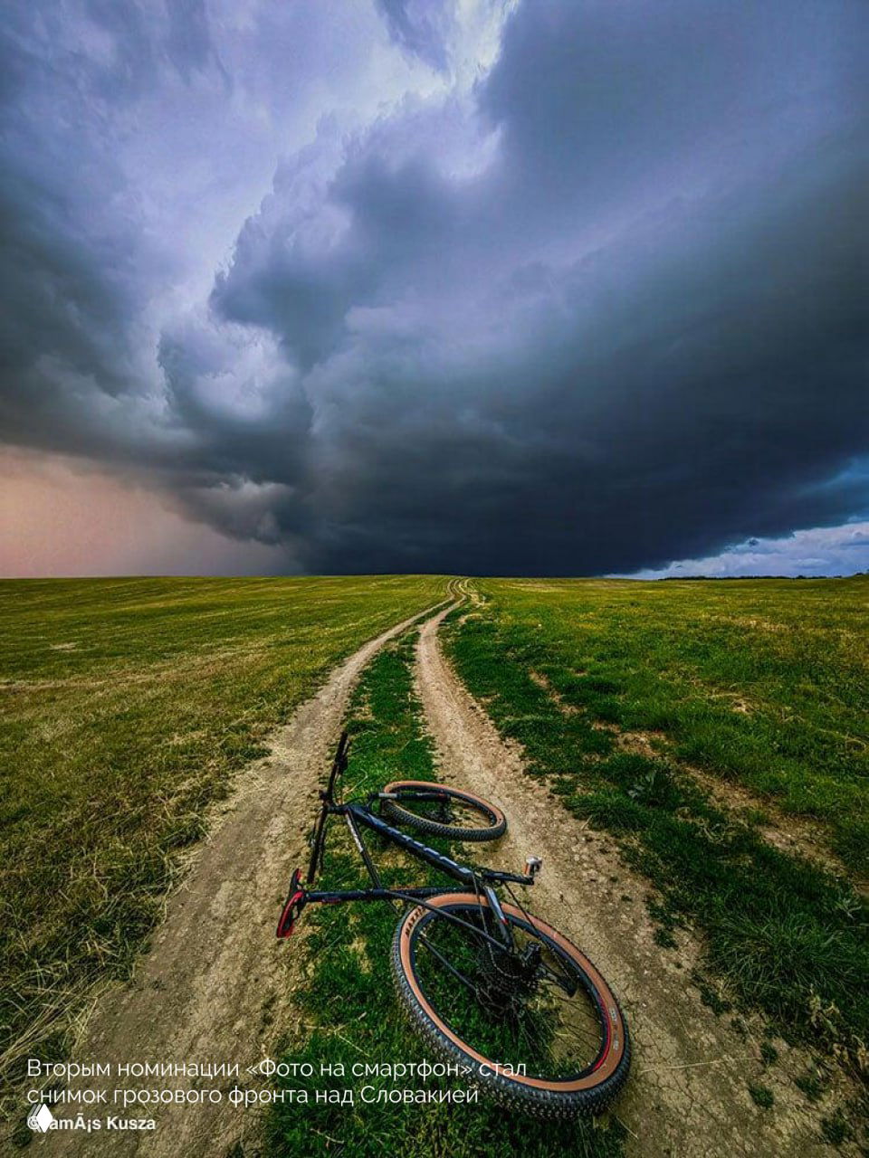 Снимок с земли: велосипед на просёлочной дороге на фоне грозового фронта над равниной, тёмные массивные облака над горизонтом.