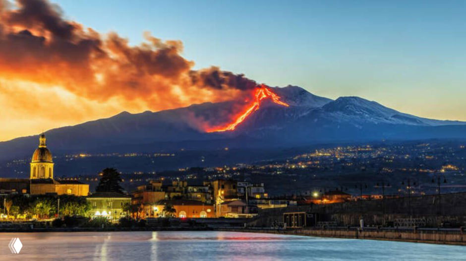 Широкий вид на Этну при закате: лавовый поток по склону вулкана, дым и пепел над горой, внизу виден город и побережье.