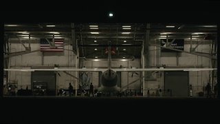 Ночной кадр из ангара: носовая часть самолета X1, технический персонал у борта и американский флаг в глубине ангарa во время подготовки к испытаниям.