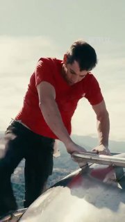 Скалолаз в красной футболке во время подъёма по высотному зданию; кадр показывает руку и корпус на фоне неба, лицо не идентифицируется.