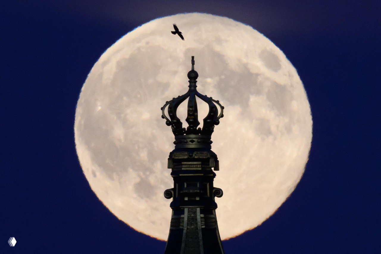 Архитектурный шпиль с короной на переднем плане и крупным полным диском Луны позади, драматичная композиция с тёмно-синим фоном
