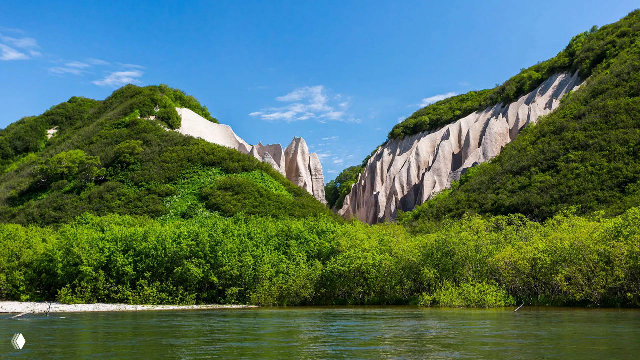 Вид с воды на белые скальные образования Кутхины Баты между зелёными склонами и спокойной поверхностью реки или залива на переднем плане.
