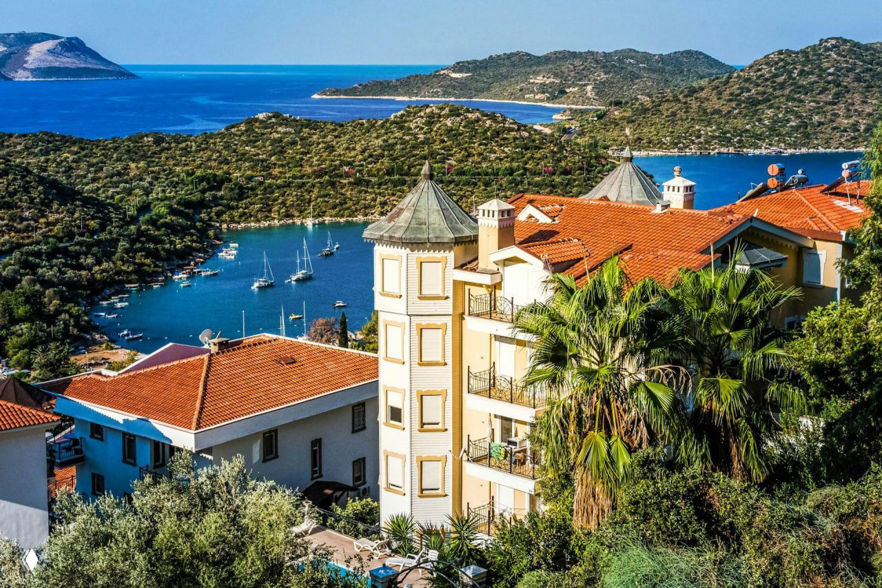 Панорама домов с красными крышами, пальм и зелёных склонов над бухтой Каша; в воде видны пришвартованные яхты и острова на горизонте.