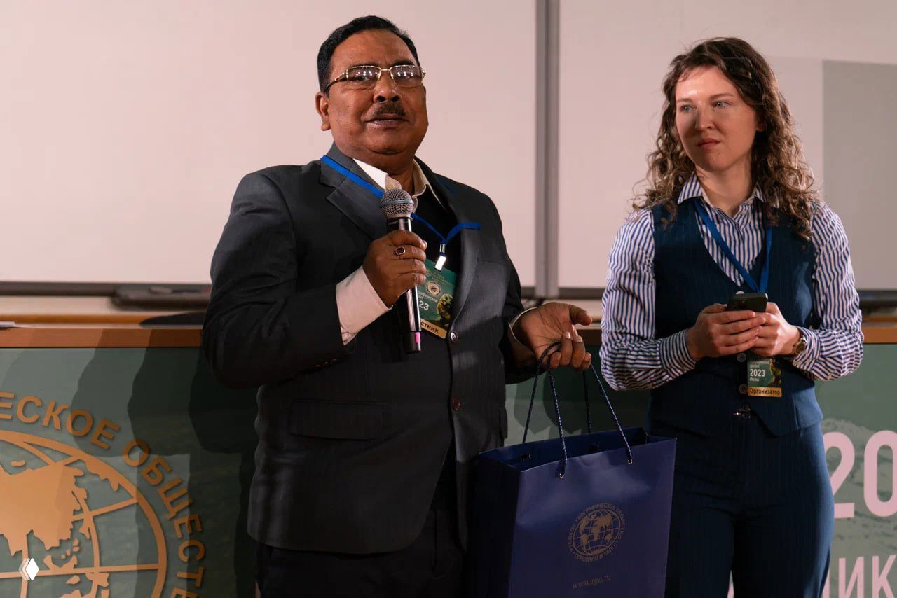 Спикер держит микрофон на сцене перед аудиторией; рядом стоит женщина с бейджем и смартфоном на фоне оформления РГО.