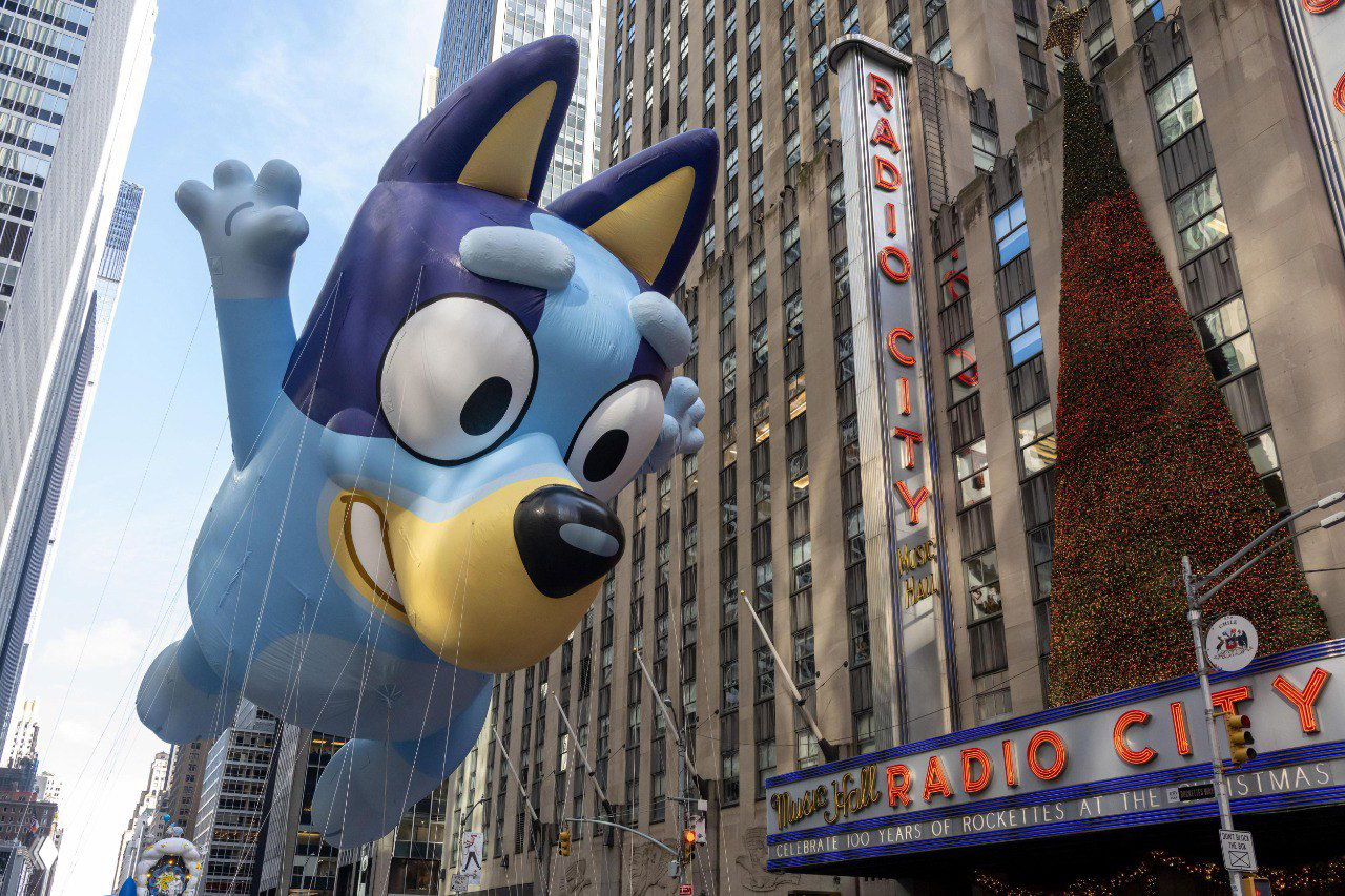 Синий шар в образе мультсобаки проходит мимо радио-театра Radio City, держатели в цветной форме ведут фигуру вдоль маршрута.