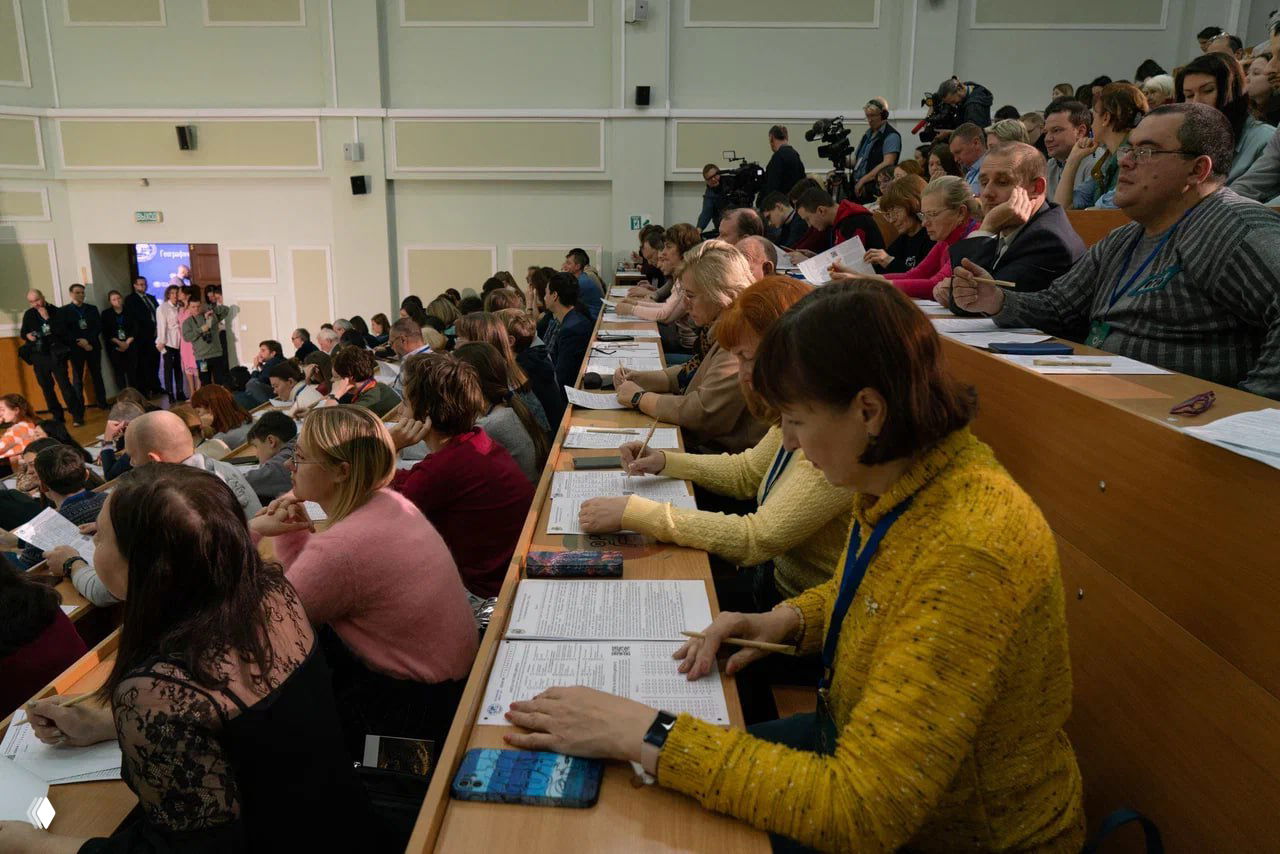 Широкий план аудитории: сотни участников на очной площадке пишут диктант за рядами столов, видно раздаточные бланки.