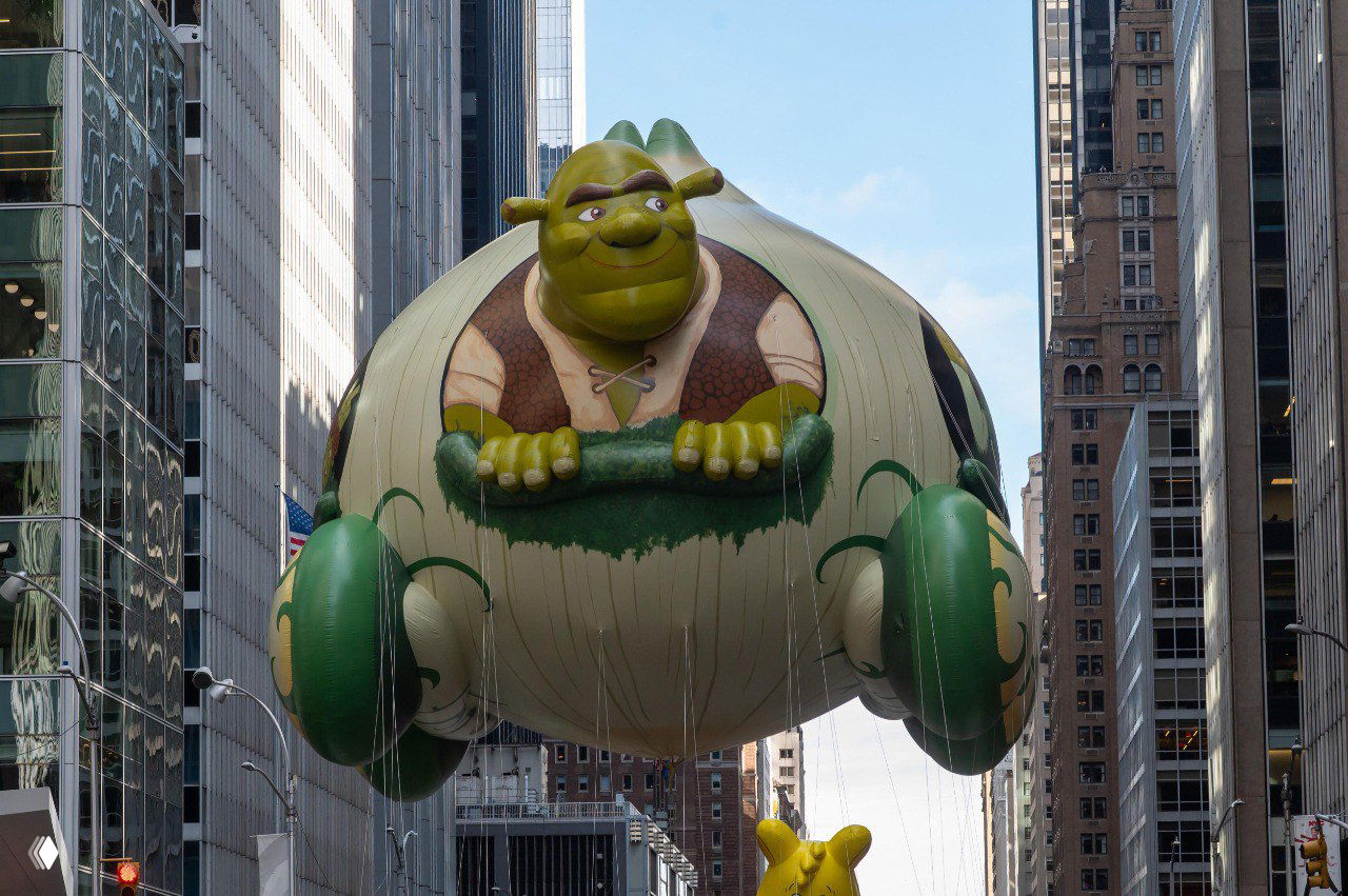 Большой надувной шар в виде Шрека парит между зданиями Манхэттена на параде Macy’s; в кадре городская архитектура и прохожие.