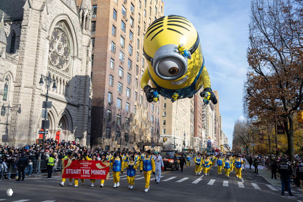 Миньон в виде огромного надувного шара пролетает над улицей, зрители заполняют бордюры, участники парада идут в униформе.