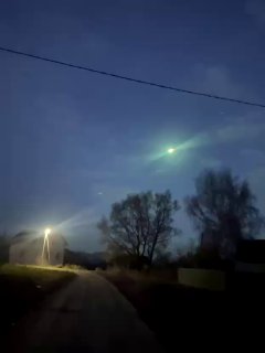 Ночное фото болида над деревней: зелёное светящееся тело в небе над дорогой, видны деревья и уличный фонарь, кадр очевидца.