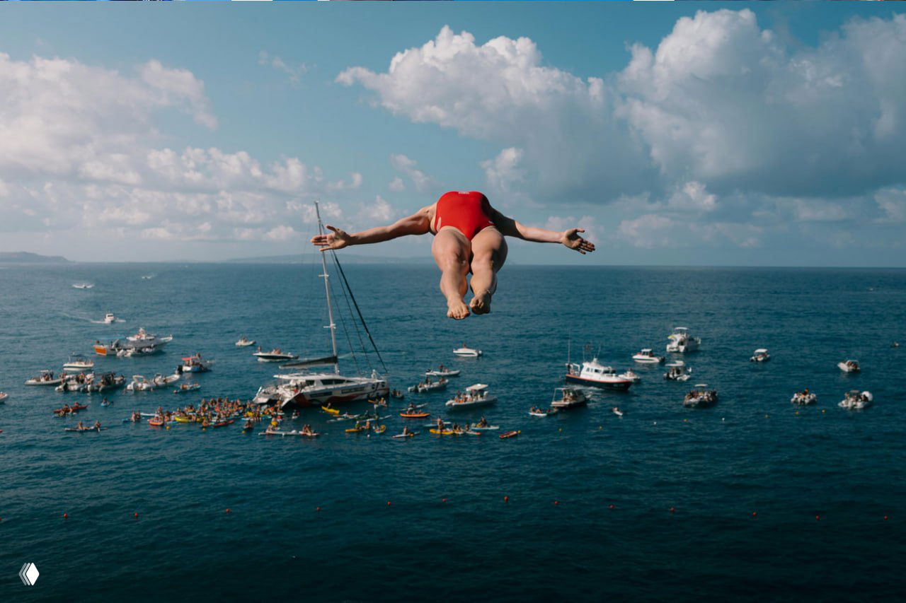 Прыгунья, отталкивающаяся над морем, на заднем плане множество лодок и зрителей — крупный атмосферный кадр с соревнований по прыжкам в воду.
