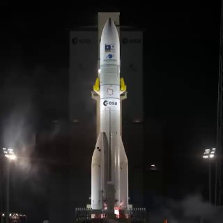 Ночная фотография ракеты Ariane 6 на стартовом комплексе: вертикальная ракета в свете прожекторов и клубы пара перед запуском, вид спереди.
